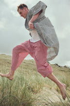 A man running barefoot through grassy dunes, wearing a black and white zigzag-patterned robe, a white Tshirt, and red striped harem pants.