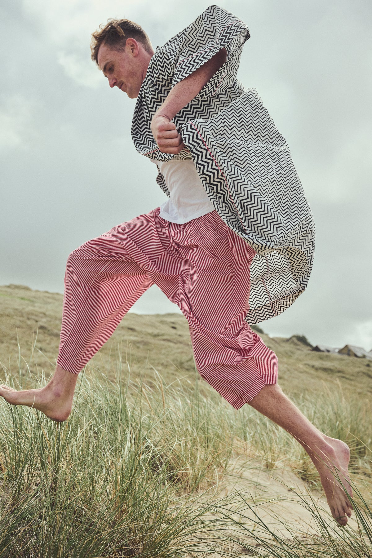 A man running barefoot through grassy dunes, wearing a black and white zigzag-patterned robe, a white Tshirt, and red striped harem pants.