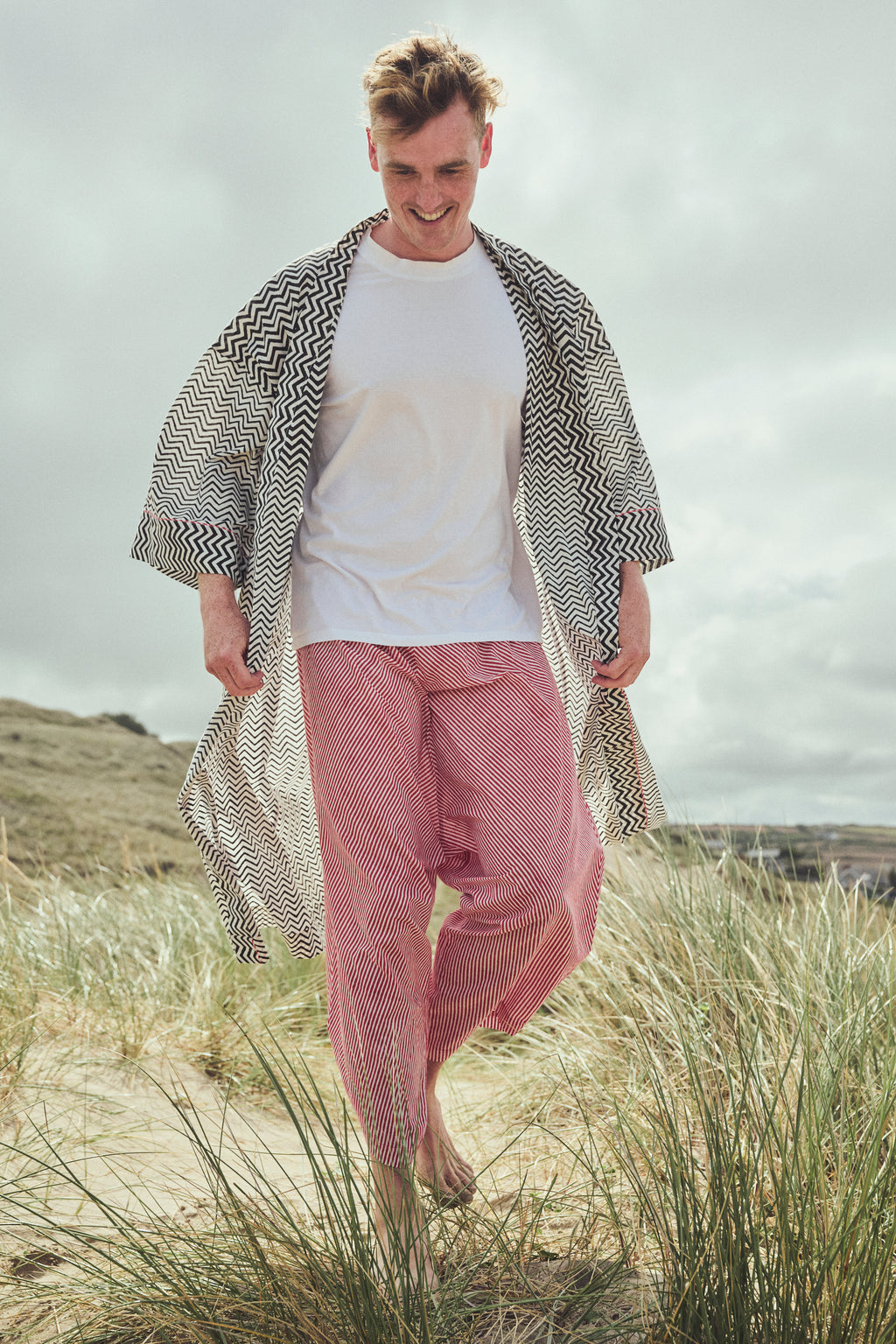 A man walking barefoot through beach grass, wearing a white t-shirt, red striped pants, and a black-and-white zigzag open-front cover-up.