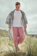 A man walking barefoot through beach grass, wearing a white t-shirt, red striped pants, and a black-and-white zigzag open-front cover-up.