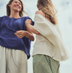 Two women outdoors wearing loose, lightweight tops and striped pants, captured from a low angle against a bright sky. One top is indigo blue and the other is ivory, paired with striped green and white bottoms.