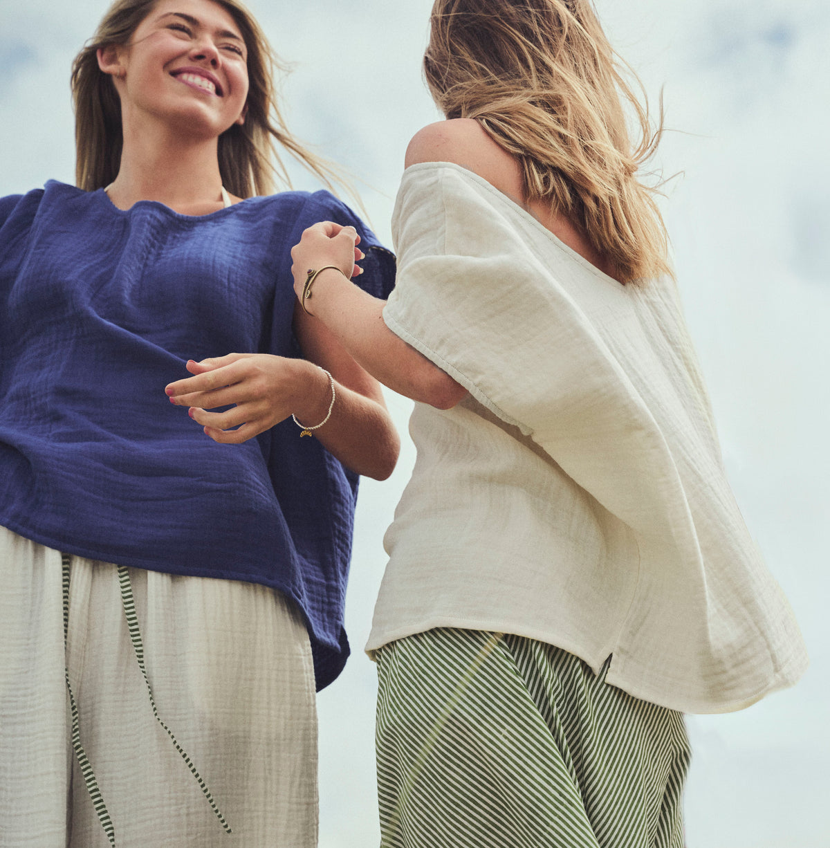 Two women outdoors wearing loose, lightweight tops and striped pants, captured from a low angle against a bright sky. One top is indigo blue and the other is ivory, paired with striped green and white bottoms.