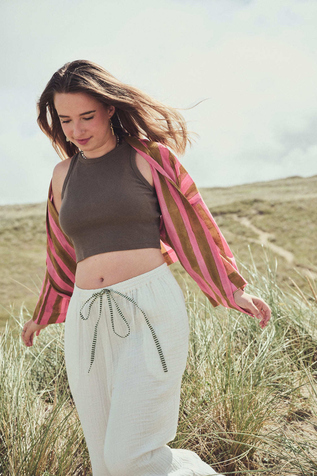 A woman outdoors in a grassy field wearing a khaki crop top, white drawstring pants, and a pink and brown striped kimono style jacket.