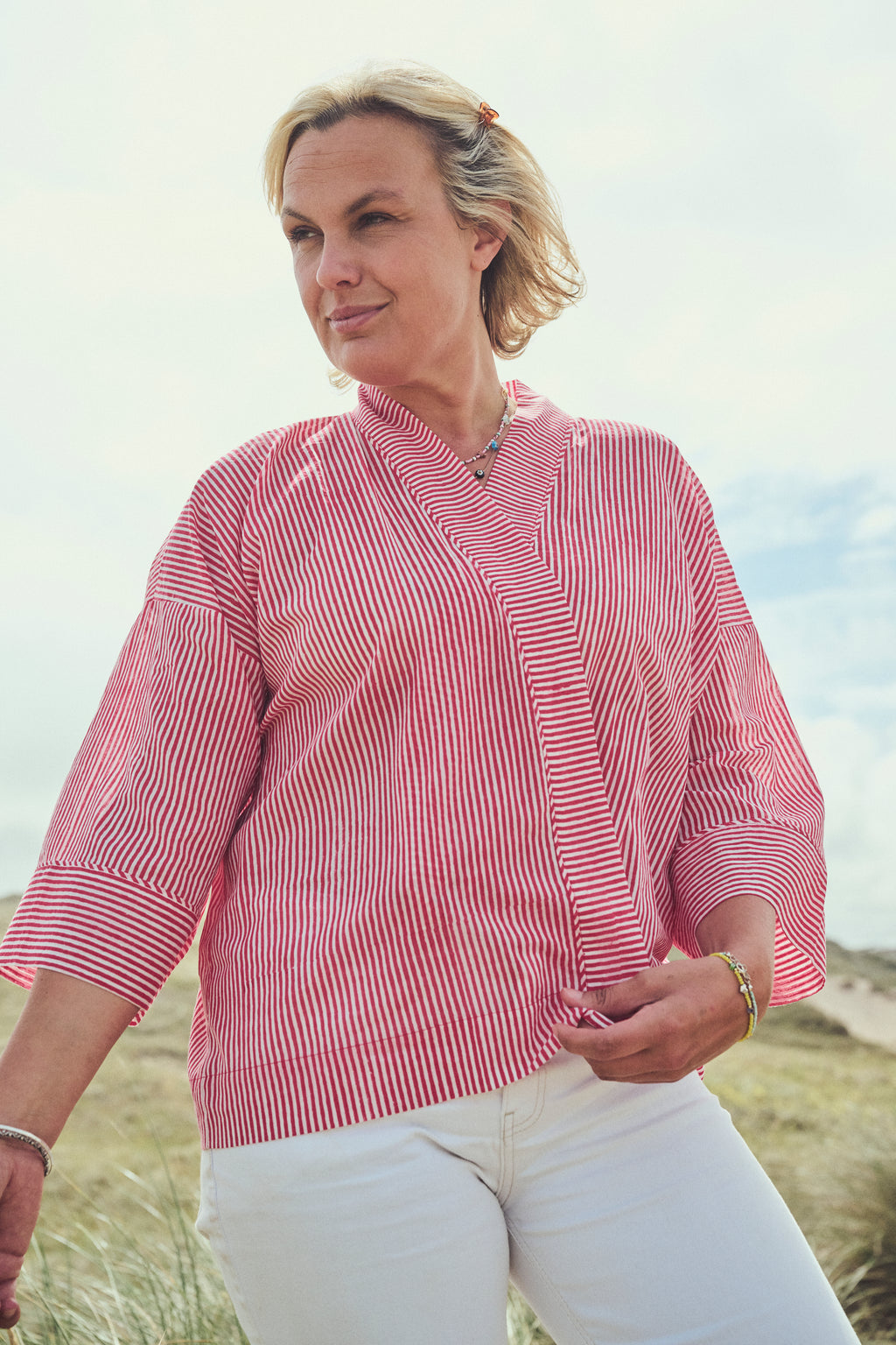 A woman wearing a red and white vertically striped wrap-style jacket with three-quarter sleeves, paired with white pants, standing outdoors in a grassy sand-dunes under a bright sky.