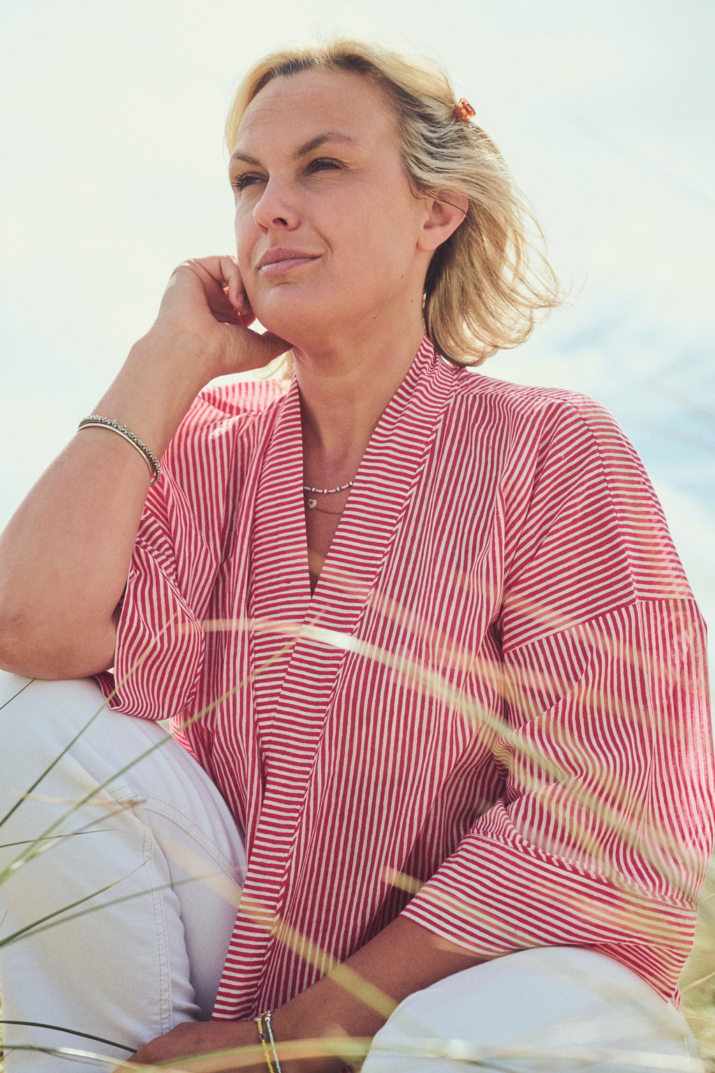 A woman with blonde hair sitting outdoors in dune grass, wearing a red and white vertically striped wrap-style top/jacket  and white pants.