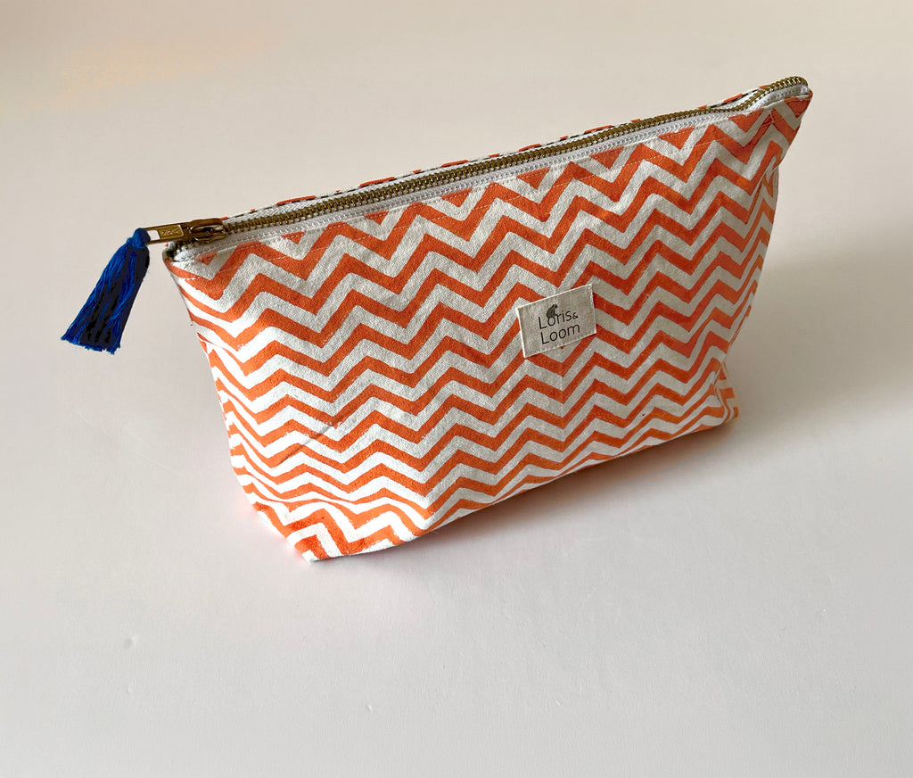 A zippered washbag with an orange and white chevron pattern, featuring a small blue tassel zipper pull and a white brand label that reads 'Loris & Loom'.